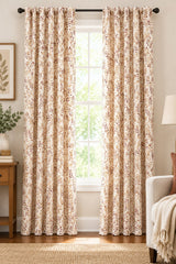 A pair of floor-length curtain panels with a beige and brown floral pattern, hanging on a black rod with matching grommets in front of a tall window.