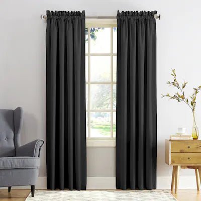 A pair of black curtain panels hanging on a silver rod over a window in a bright room, with a gray armchair on the left and a wooden side table with a vase on the right.