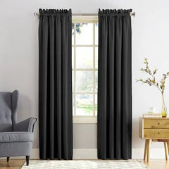 A pair of black curtain panels hanging on a silver rod over a window in a bright room, with a gray armchair on the left and a wooden side table with a vase on the right.